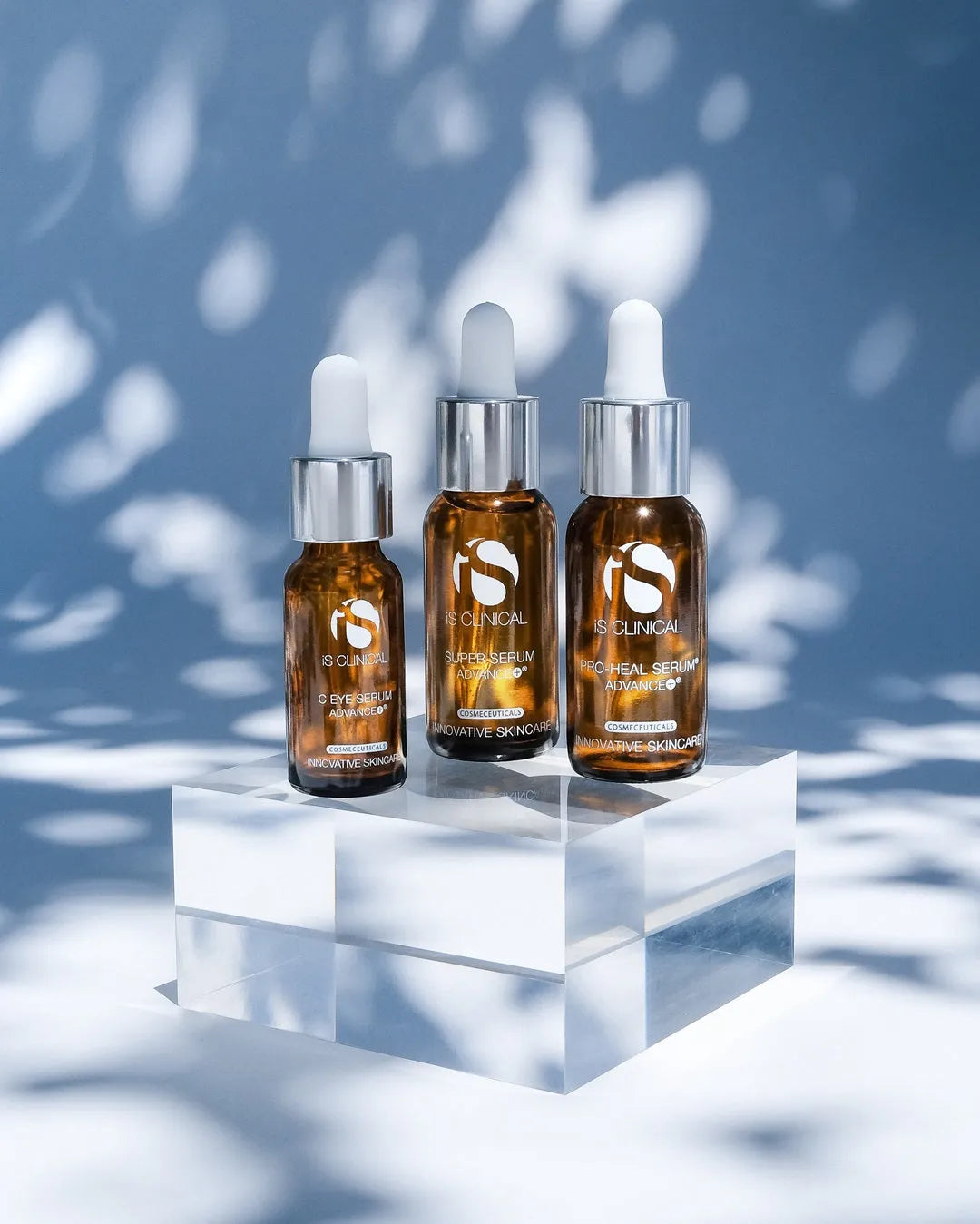 Three amber glass dropper bottles of iS Clinical serums on a clear acrylic block with dappled light background