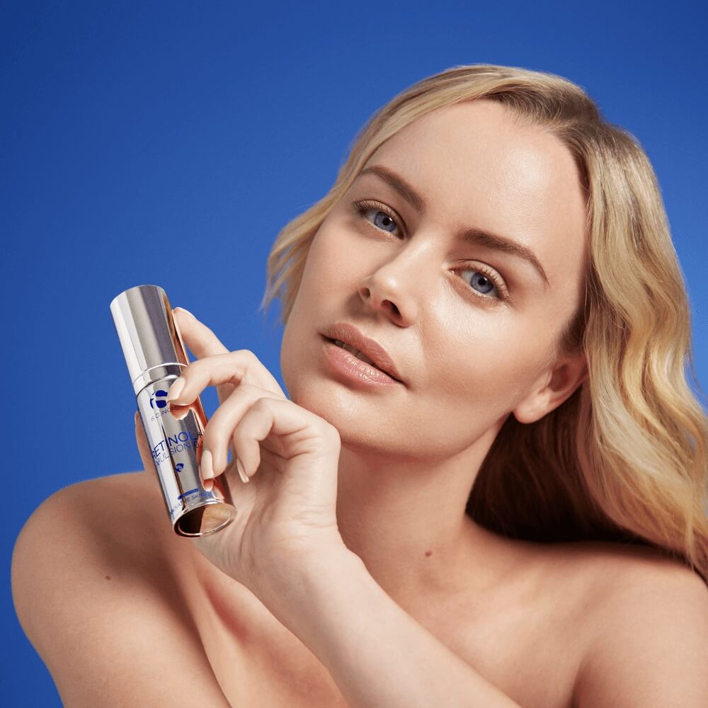 Woman with clear skin holding a silver bottle of iS Clinical Retinol Emulsion against blue background