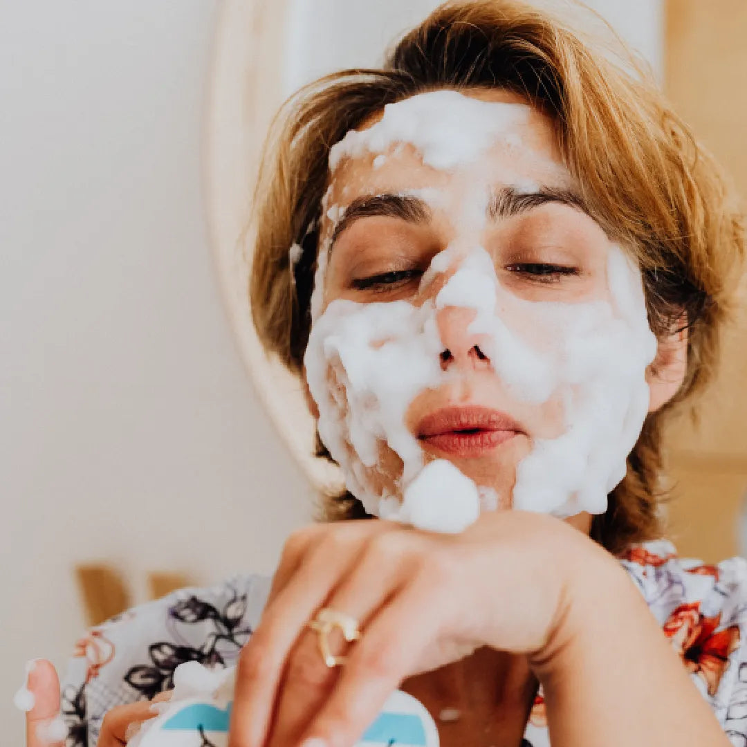 Person with foaming white facial cleanser applied on face, holding product container