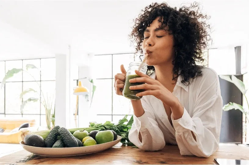 Person with curly hair in white shirt drinking green smoothie at wooden table with plate of green vegetables and fruits