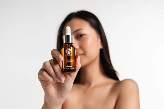 Hand holding amber glass dropper bottle of iS Clinical Pro-Heal Serum Advance+ with blurred person in background