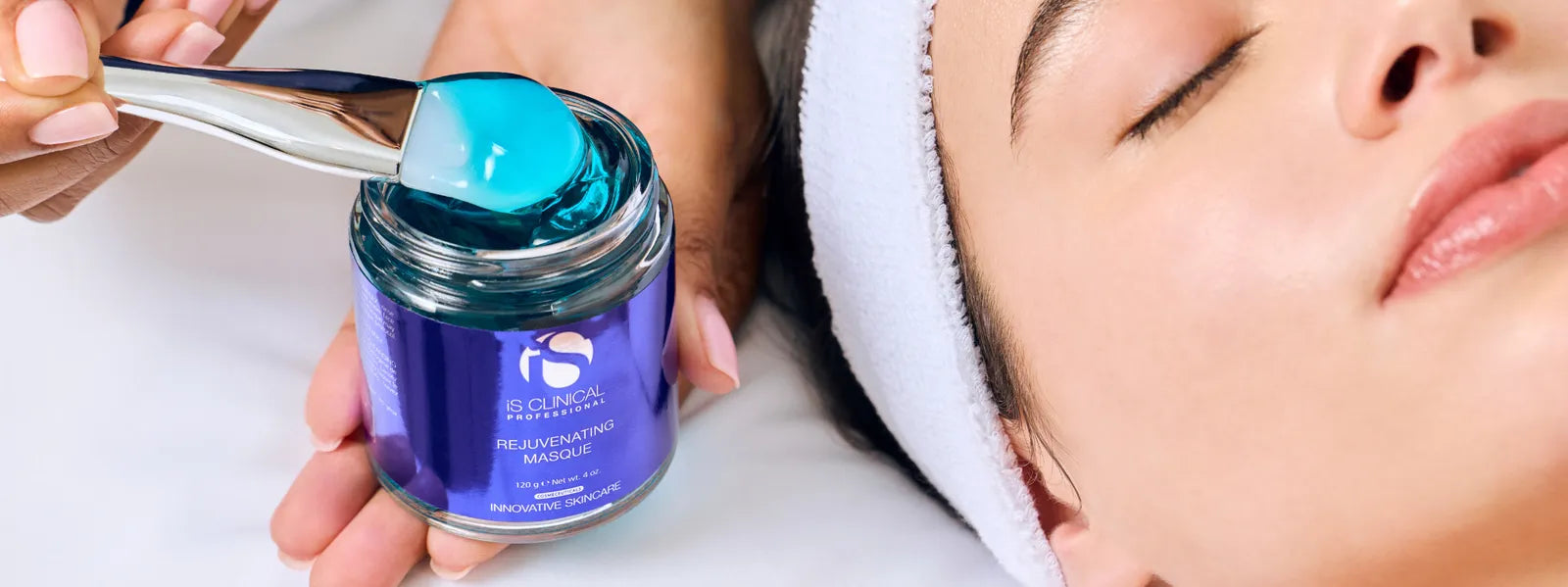 Hand holding iS Clinical Rejuvenating Masque jar with blue gel and brush near relaxed face with white headband