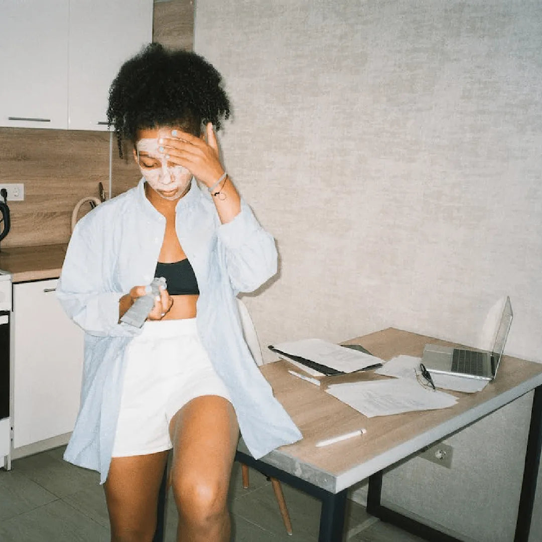 Person applying white facial cream while sitting on kitchen table near laptop and papers