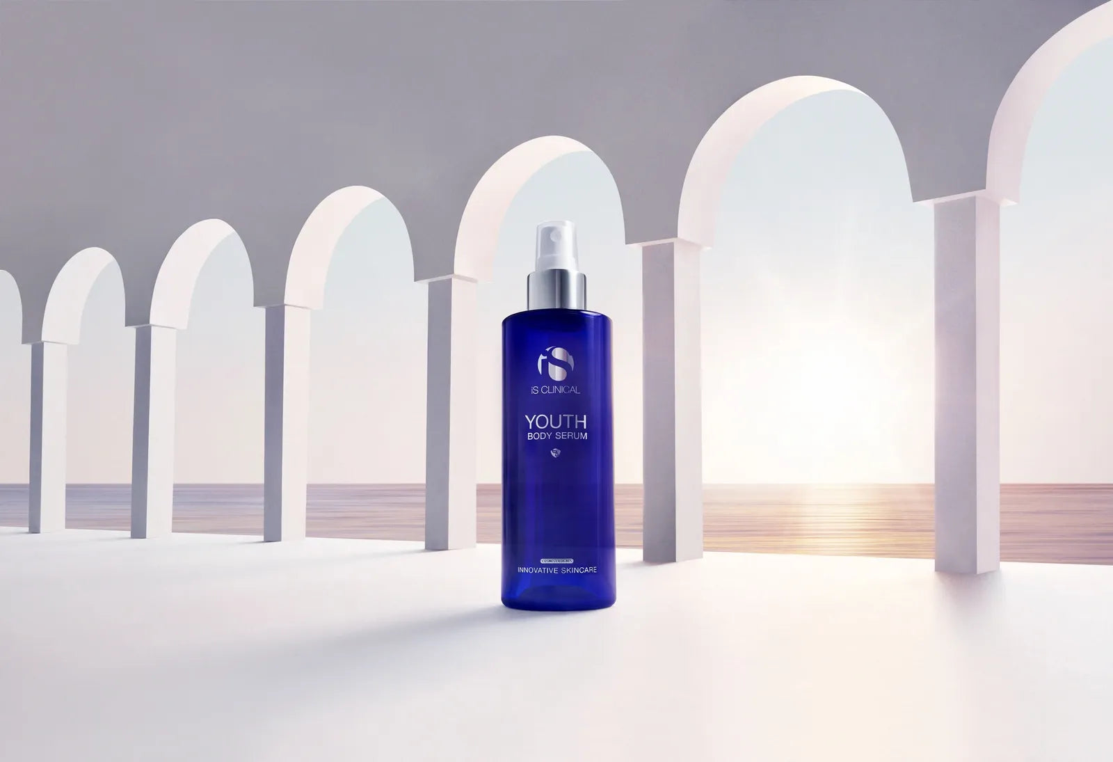 Blue iS Clinical Youth Body Serum bottle with silver spray cap against white arches and ocean background