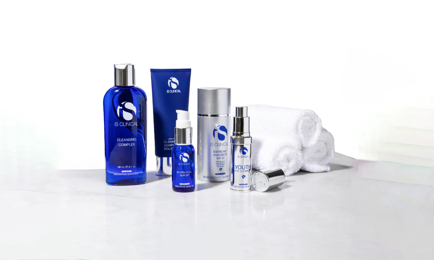 iS Clinical skincare products including Cleansing Complex, Hydra-Cool Serum, Extreme Protect SPF 40, Youth Eye Complex, and white rolled towels on a white surface
