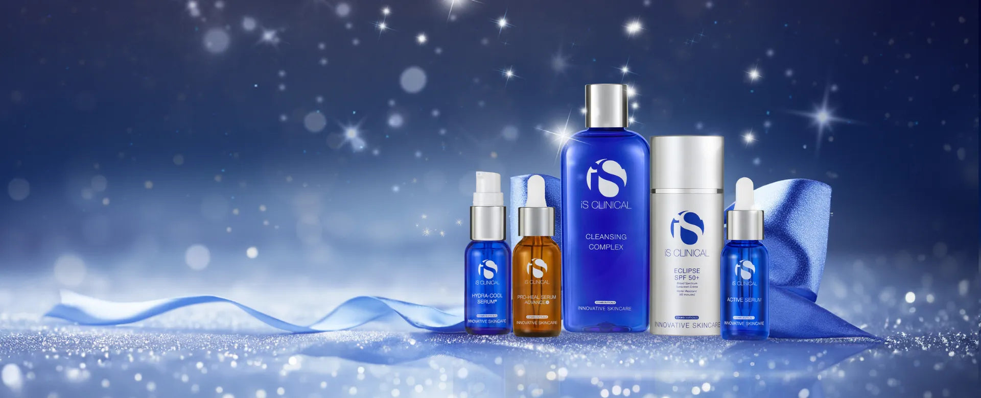 iS Clinical skincare collection with blue and amber bottles, including Cleansing Complex and Active Serum, on sparkling surface with blue ribbon and starry background