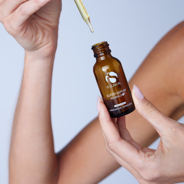 Hand holding amber iS Clinical Super Serum Advance+ bottle with dropper releasing serum drop