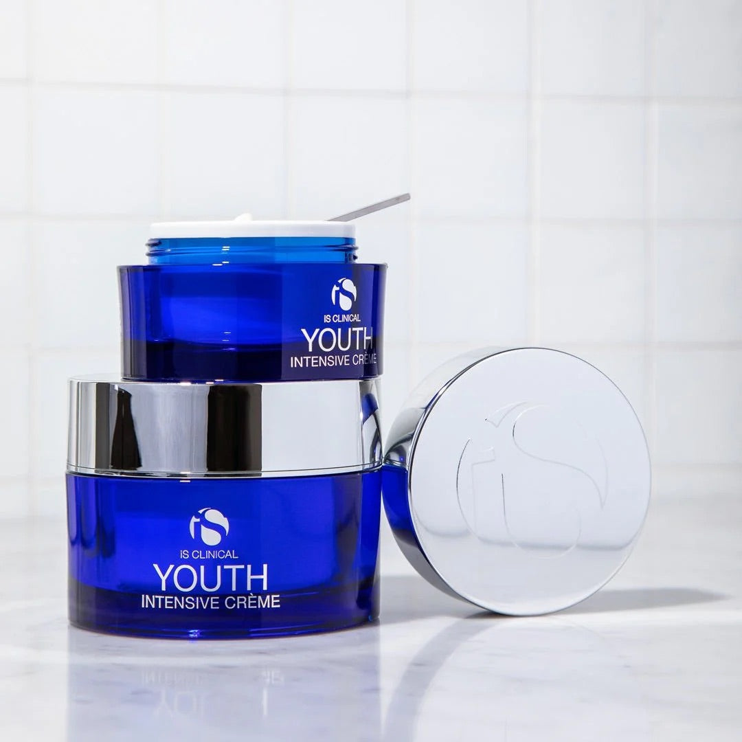 Two blue jars of iS Clinical Youth Intensive Crème with silver lids on a white surface, one jar open with cream and a small spatula