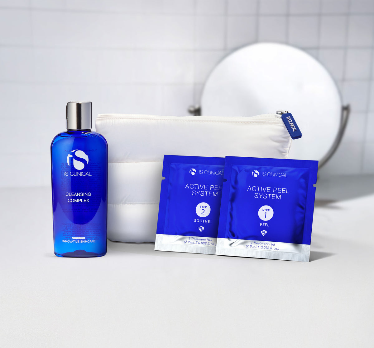 iS Clinical Cleansing Complex bottle and two Active Peel System treatment pad packets beside white zip pouch on white surface