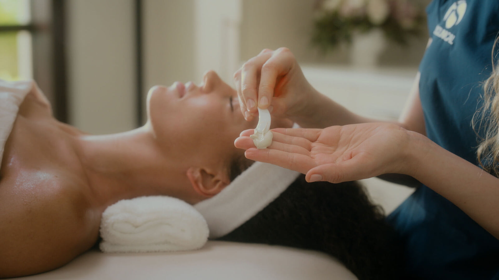 Close-up of hands holding white cream with a woman lying down wearing a white headband