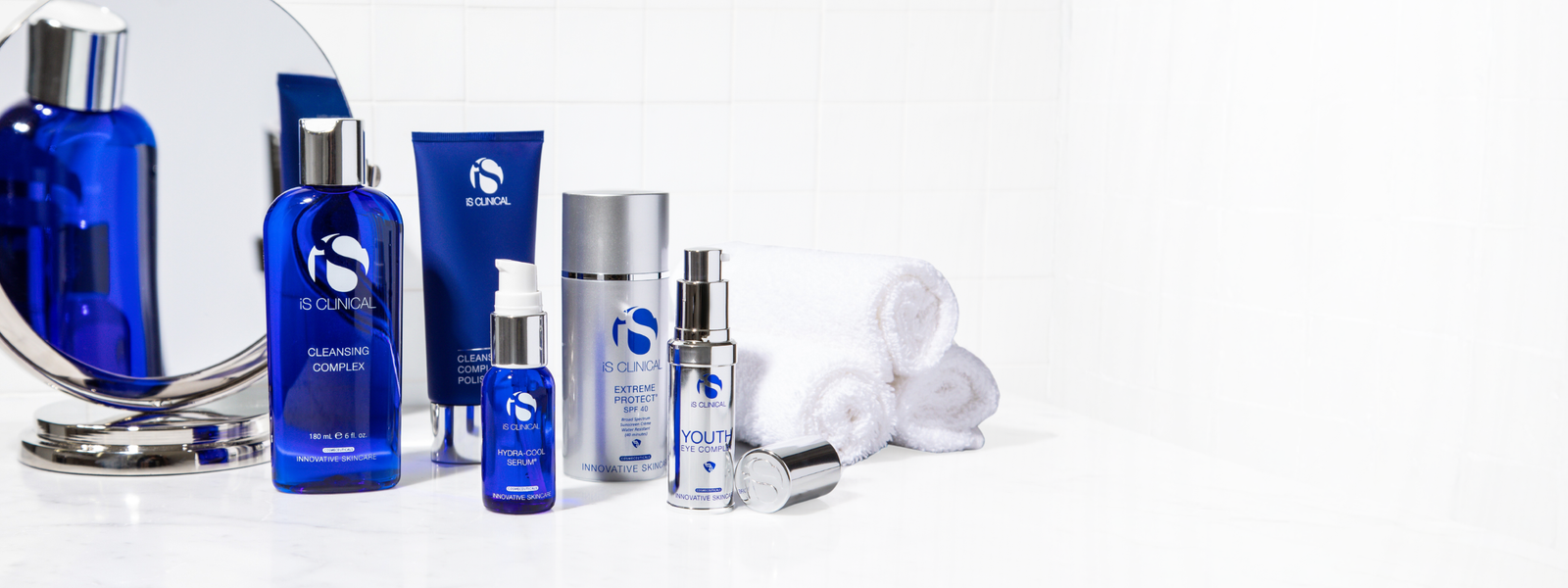 iS Clinical skincare products including Cleansing Complex, Hydra-Cool Serum, Extreme Protect SPF 40, and Youth Eye Complex with rolled white towels and round mirror