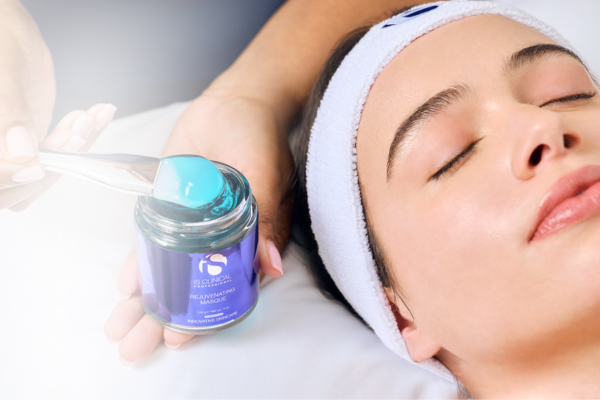 Close-up of a woman with eyes closed wearing a white headband, a hand holding iS Clinical Rejuvenating Masque jar with blue gel and spatula