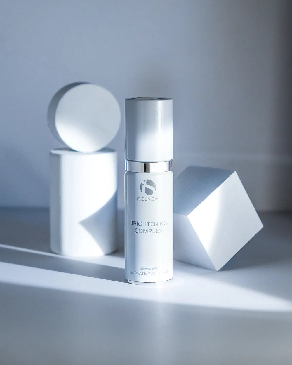iS Clinical Brightening Complex bottle with silver cap against white geometric shapes on a white surface