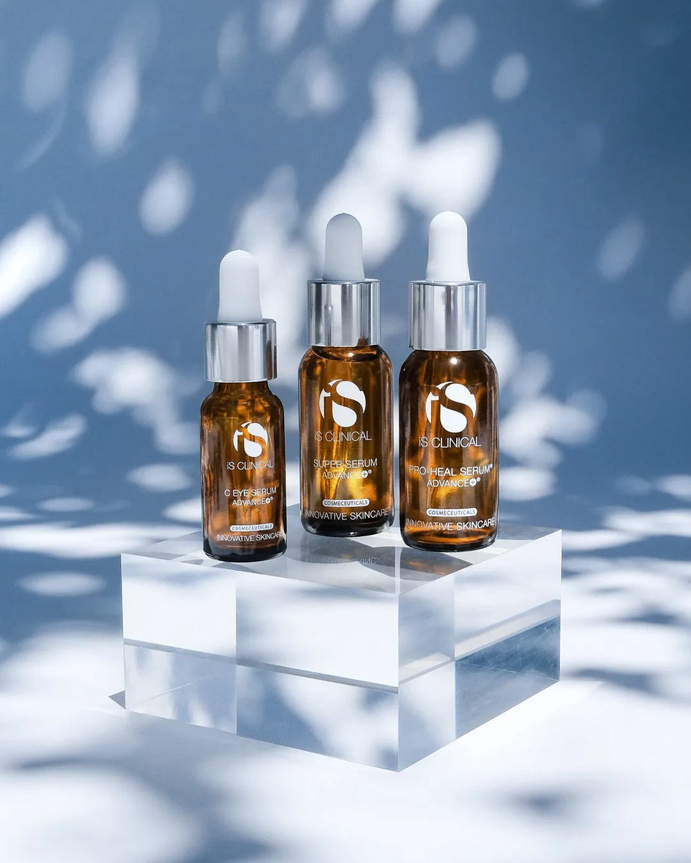 Three amber glass dropper bottles of iS Clinical serums on a clear acrylic block with dappled light background
