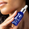 Hand holding blue glass bottle of iS Clinical GeneXC Serum with white dropper near face