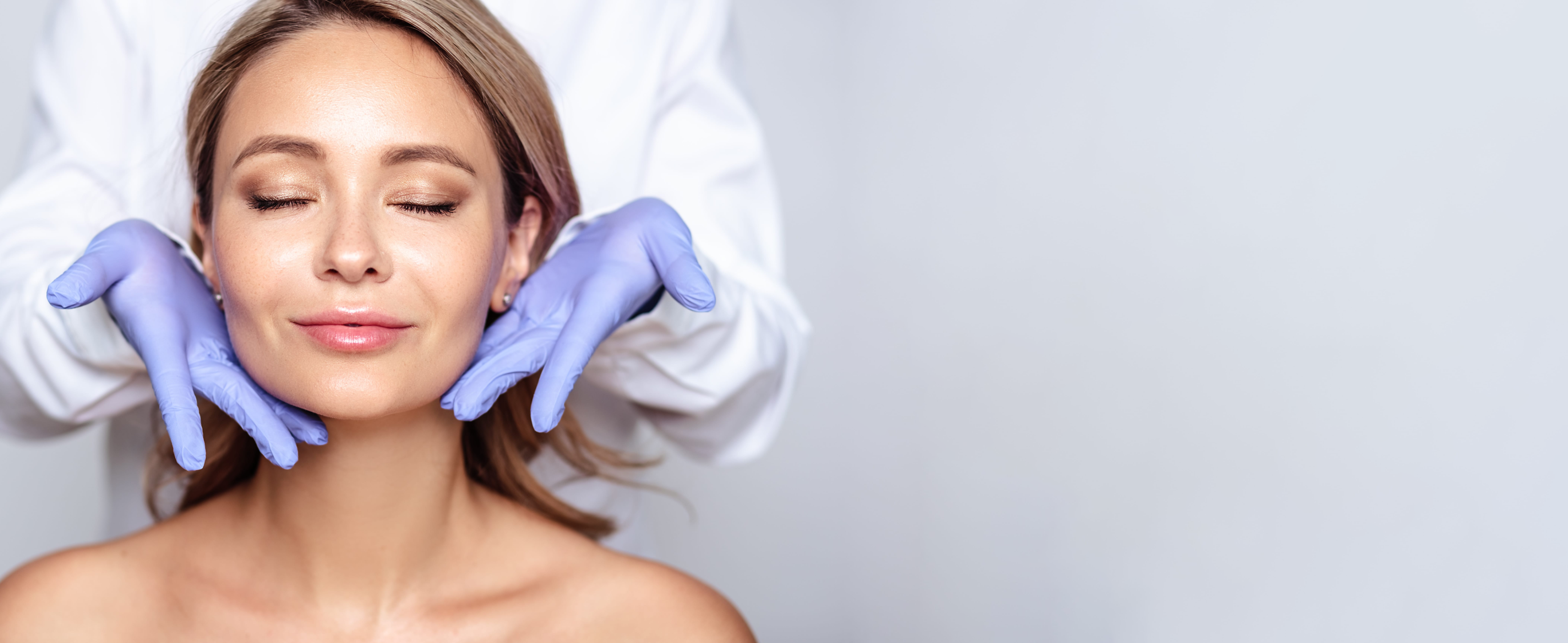 Woman with closed eyes receiving facial treatment from hands in blue gloves against white background