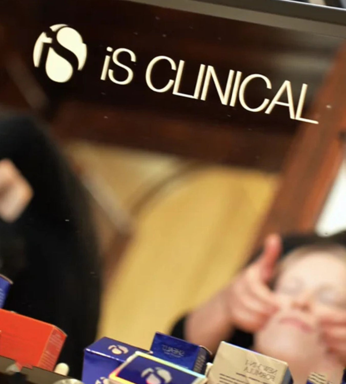 Reflection of iS Clinical logo on mirror with blurred face receiving facial massage and skincare boxes