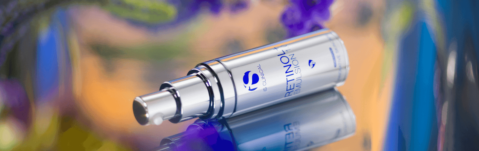 Silver pump bottle of iS Clinical Retinol+ Emulsion lying on reflective surface with blurred colourful background