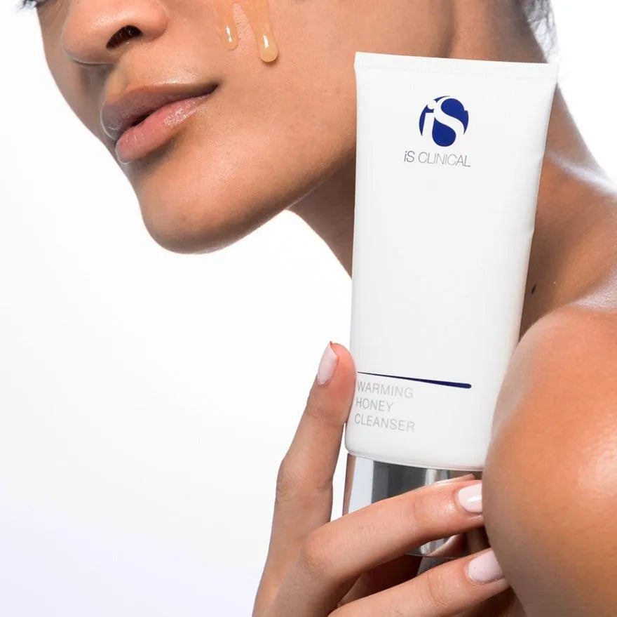 Person holding iS Clinical Warming Honey Cleanser tube with honey-coloured cleanser on cheek
