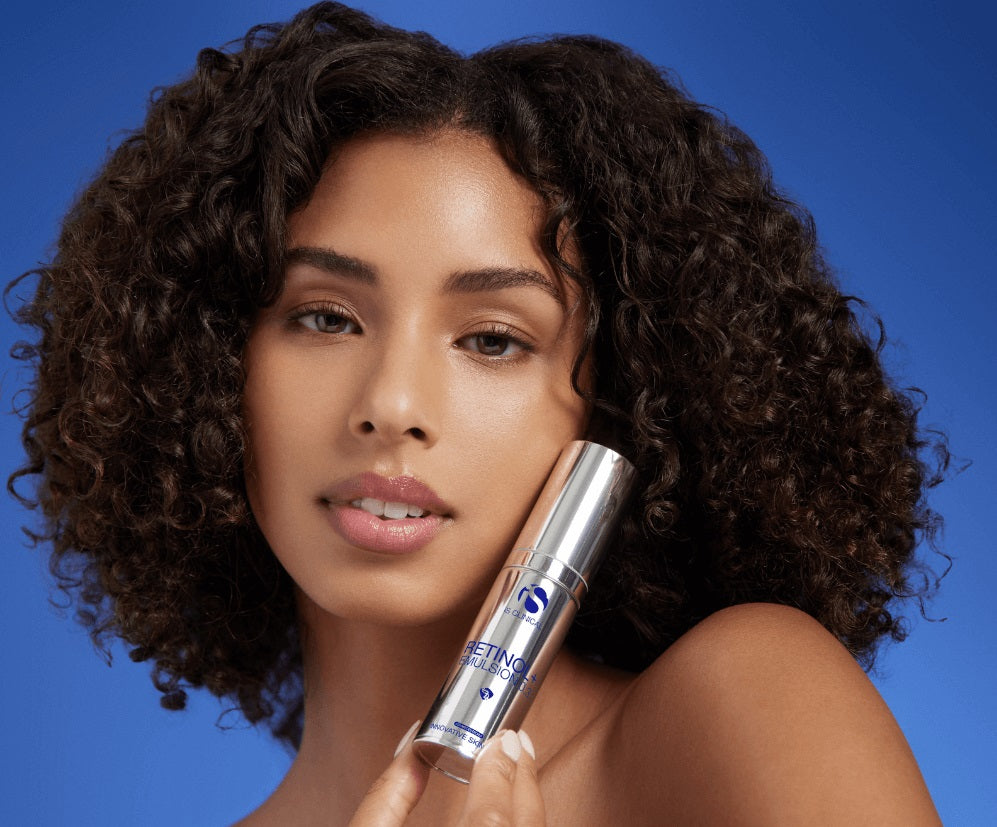 Woman with curly hair holding iS Clinical Retinol Emulsion+ bottle against blue background