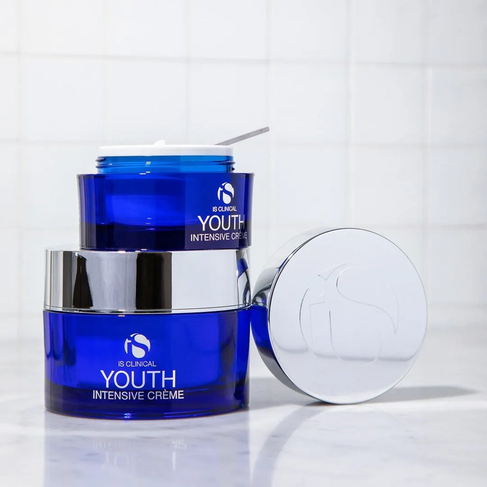 Two blue jars of iS Clinical Youth Intensive Crème with silver lids, one open showing white cream and spatula