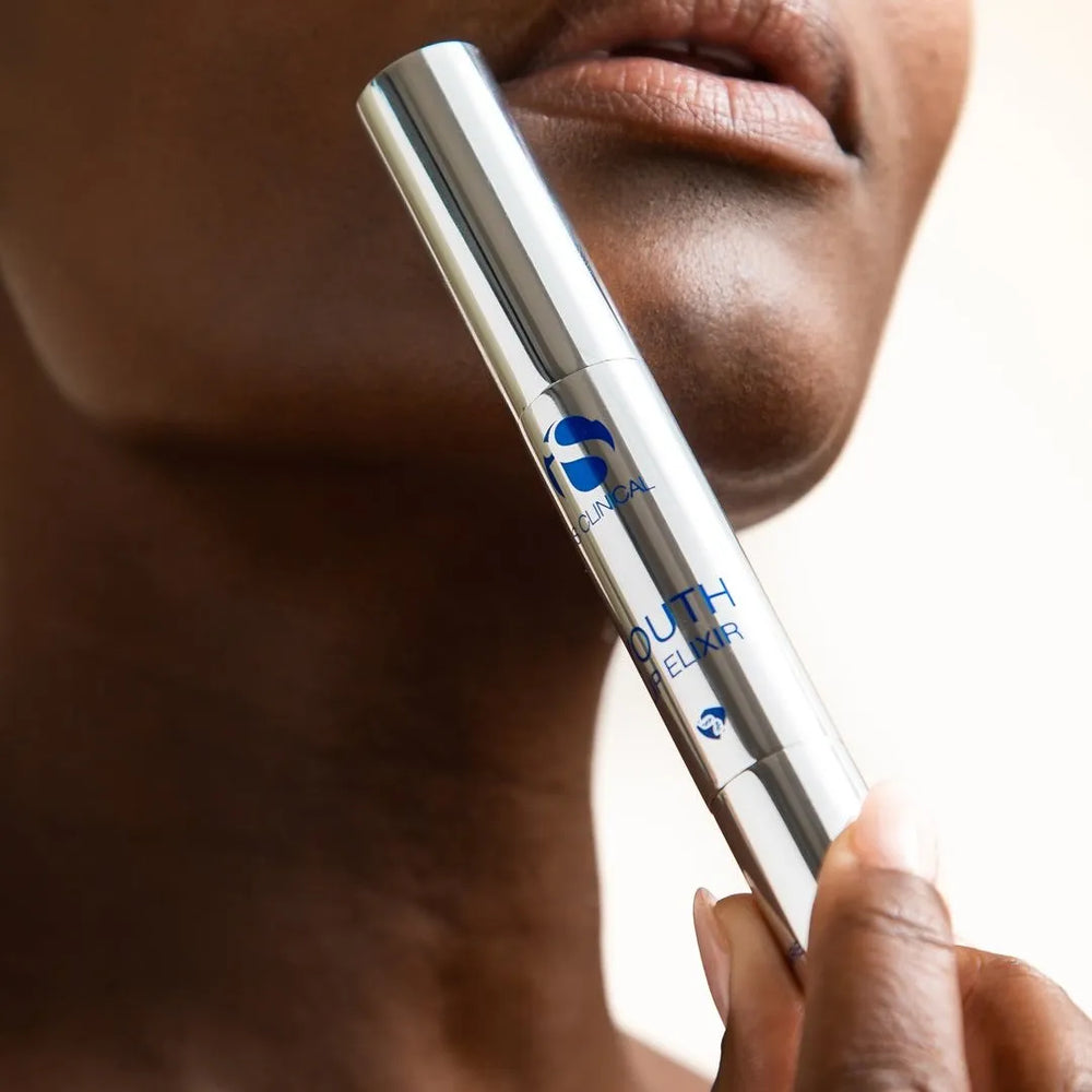 Hand holding iS Clinical Youth Lip Elixir silver tube near lips and chin on light background